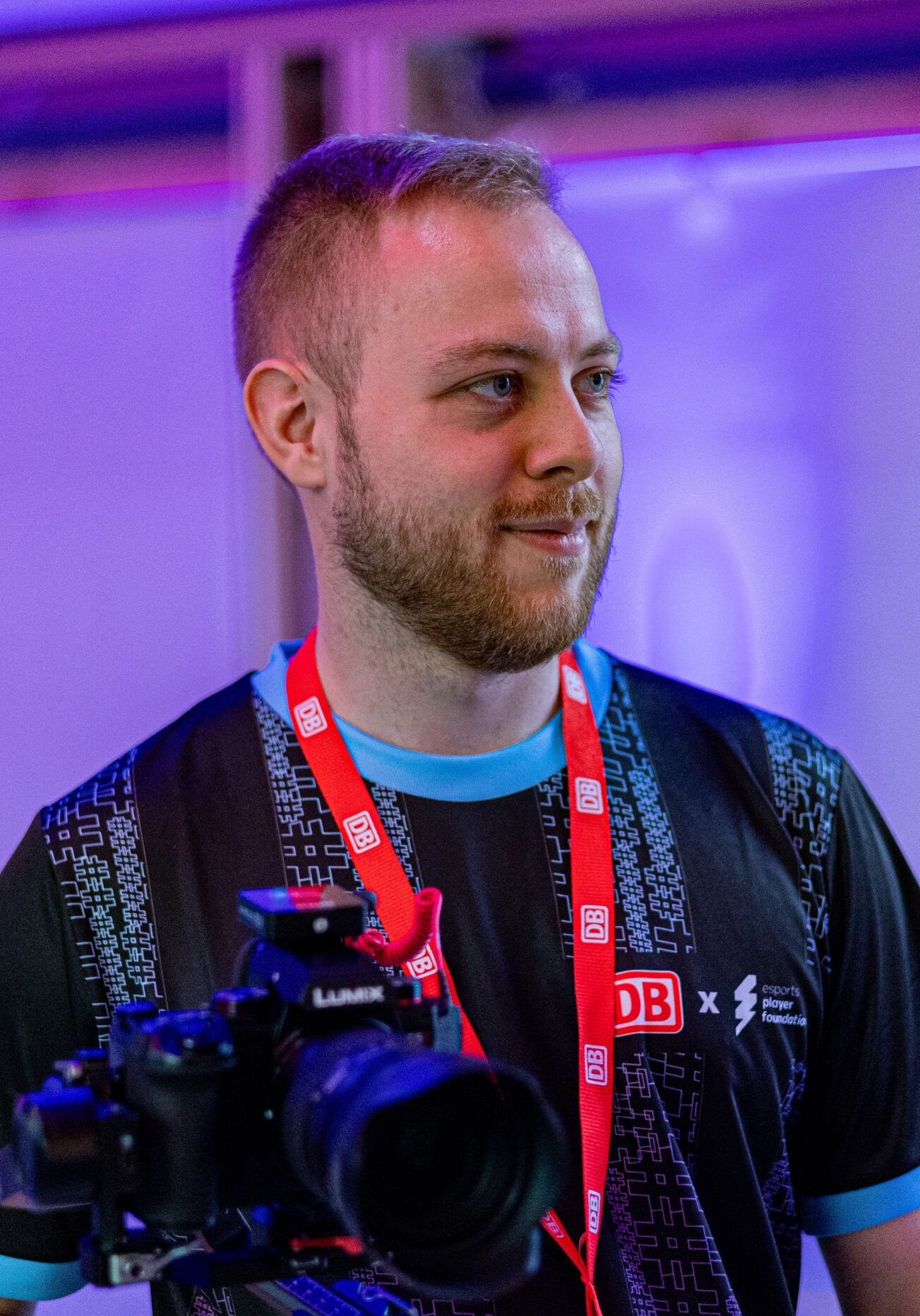Jannik Ebben Fotograf und Videograf bei der esportsplayerfoundation - Er ist bei der Arbeit zu sehen und hält in seiner rechten Hand eine Kamara mit dem Stativ. Um den Hals trägt er ein Layer mit DB Logo