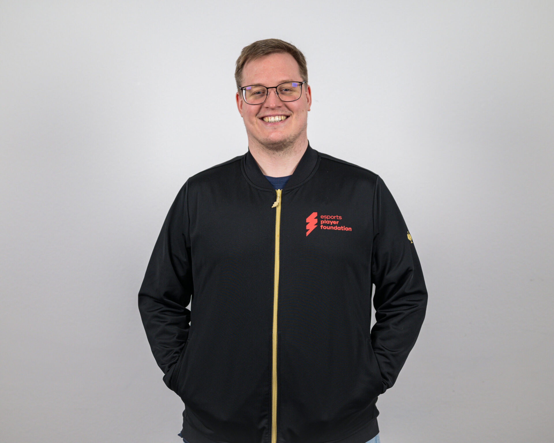 Alexander "AlexR" Frisch - Sportlicher Leiter Valorant bei der esports player foundation im Portrait - Die Hände sind seitlich in den Taschen einer Jacke. Er lacht