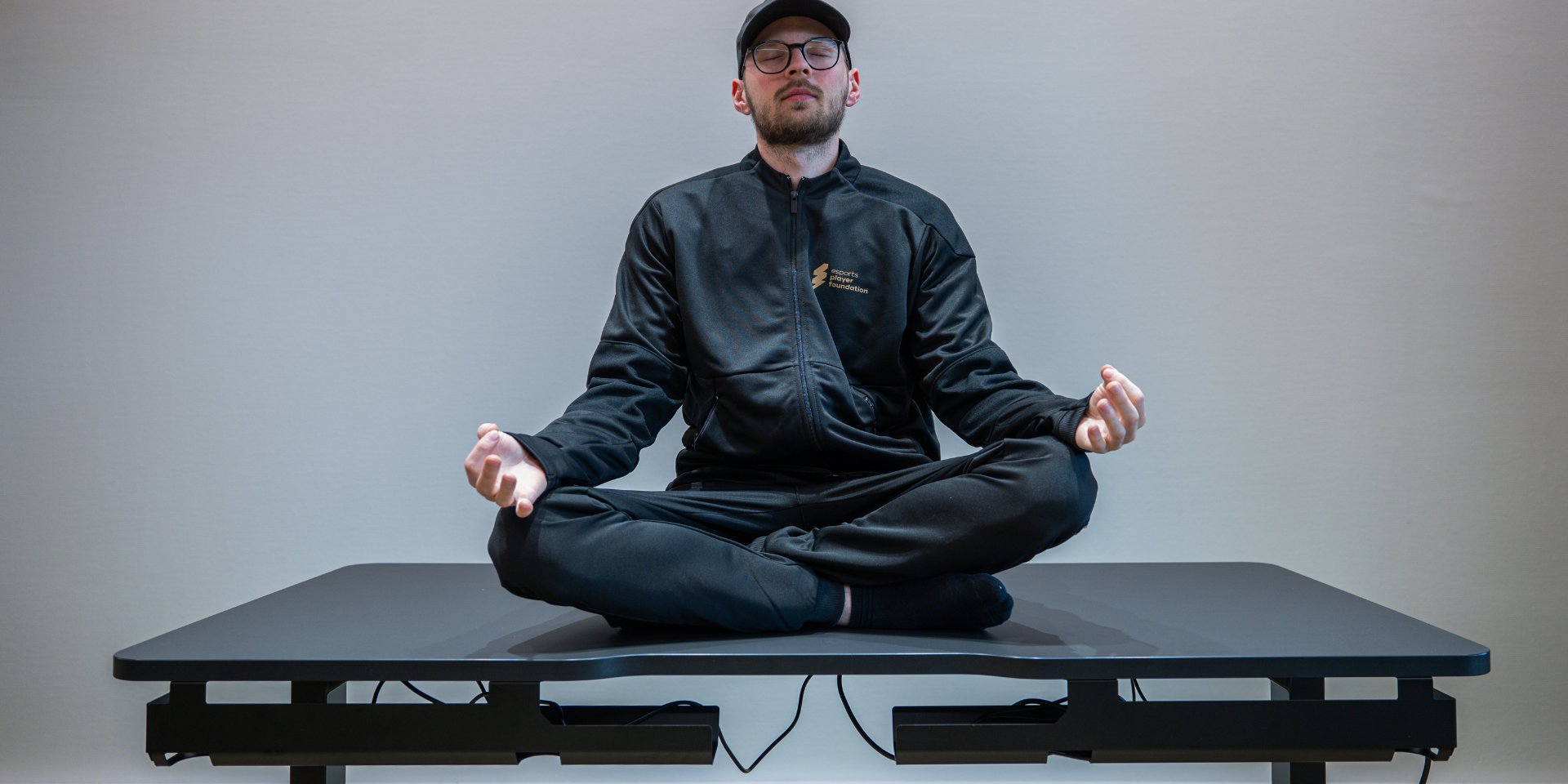 LeetDesk Gaming Tisch Esports Ein junger Mann mit Brille mit Klamotten der esports player foundation, sitzt auf einem Gaming Tisch der Marke LeetDesk. LeetDesk und die esports player foundation feiern mit diesem Bild ihre Partnerschaft.